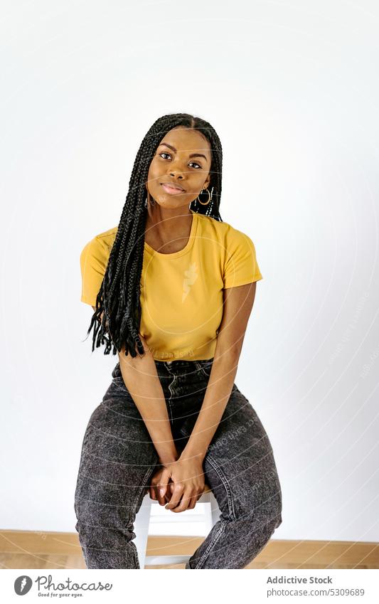Lächelnde schwarze Frau sitzt auf einem Stuhl vor einer weißen Wand positiv trendy Geflecht Inhalt Vorschein Stil selbstbewusst jung lässig ethnisch Windstille