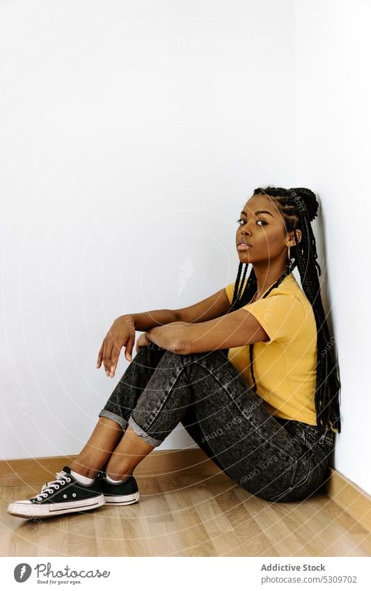 Selbstbewusste schwarze Frau sitzt auf dem Boden und schaut in die Kamera Windstille trendy ernst Stil Vorschein Wand selbstbewusst Outfit Mode jung