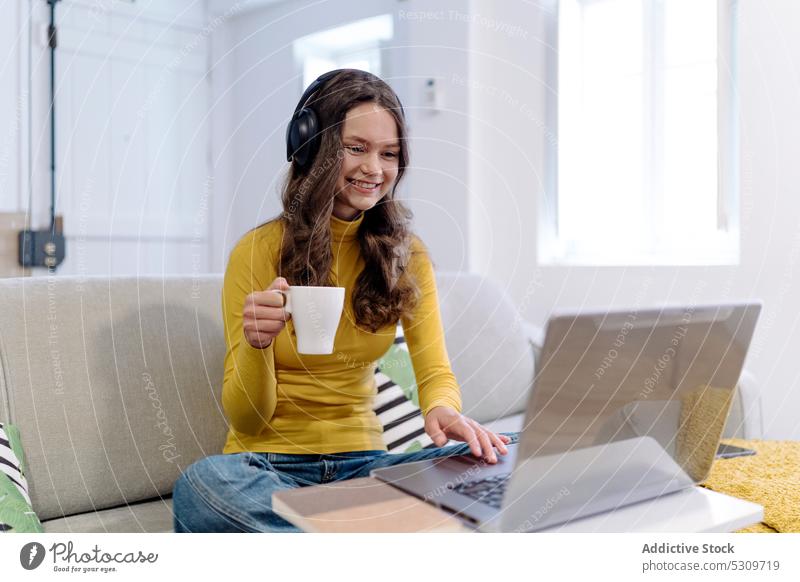 Konzentrierte Frau mit Kopfhörern und Kaffeetasse arbeitet am Laptop auf dem Sofa abgelegen freiberuflich Arbeit benutzend Telearbeit Browsen jung heimwärts