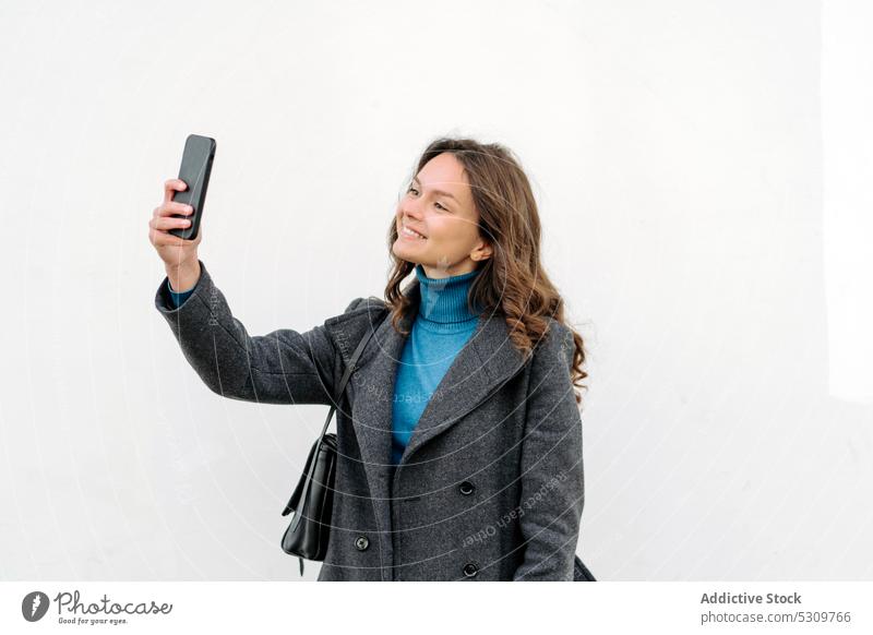 Lächelnde Frau nimmt Selfie auf Smartphone auf weißem Hintergrund benutzend Selbstportrait heiter Tasche Glück Mantel Apparatur Gerät Mobile Funktelefon
