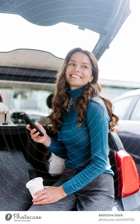 Lächelnde Frau mit Smartphone im Kofferraum eines Autos PKW Automobil Glück Kaffee Browsen Imbissbude jung Telefon Mobile Apparatur lange Haare Gerät Anschluss