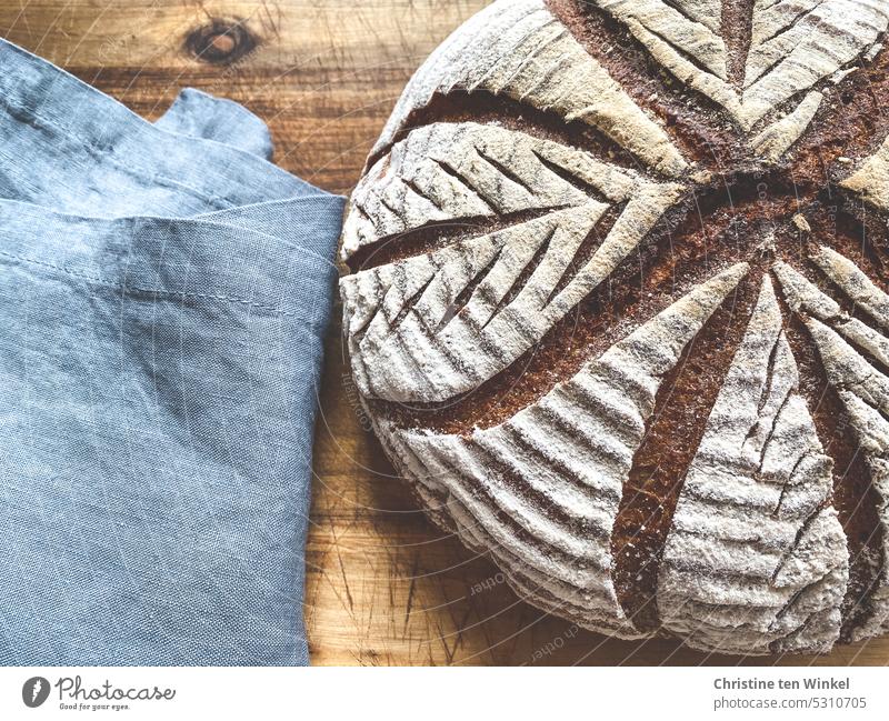 Ein selbstgebackenes Brot mit Blattmuster liegt auf einem Holzbrett neben einem blauen Leinentuch Brotmuster Kruste knusprig Brotliebe natürlich Tradition