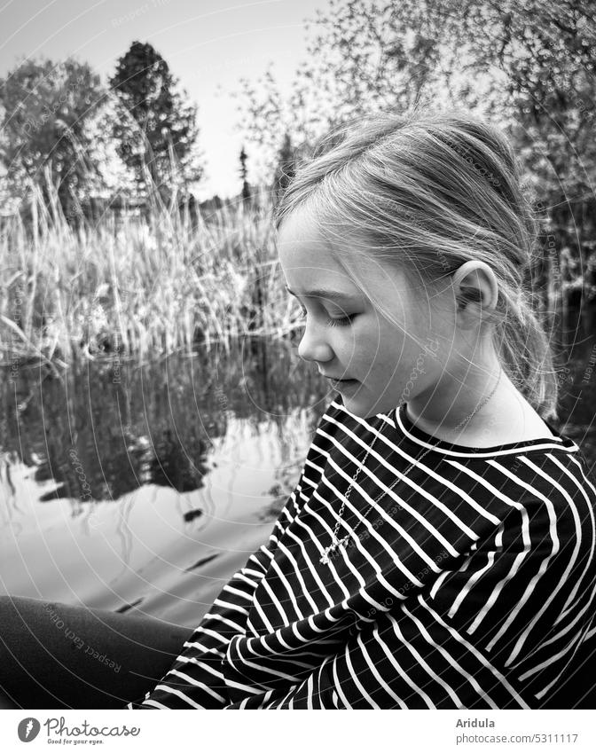 Mädchen sitzt im Tretboot auf einem See und schaut nach unten. Im Hintergrund: See und Ufer s/w