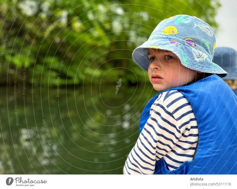 Kind sitzt in einem Boot und schaut skeptisch nach hinten auf das Wasser. Es trägt einen Sonnenhut und eine Schwimmweste Tretboot Blick unsicher Angst ängstlich