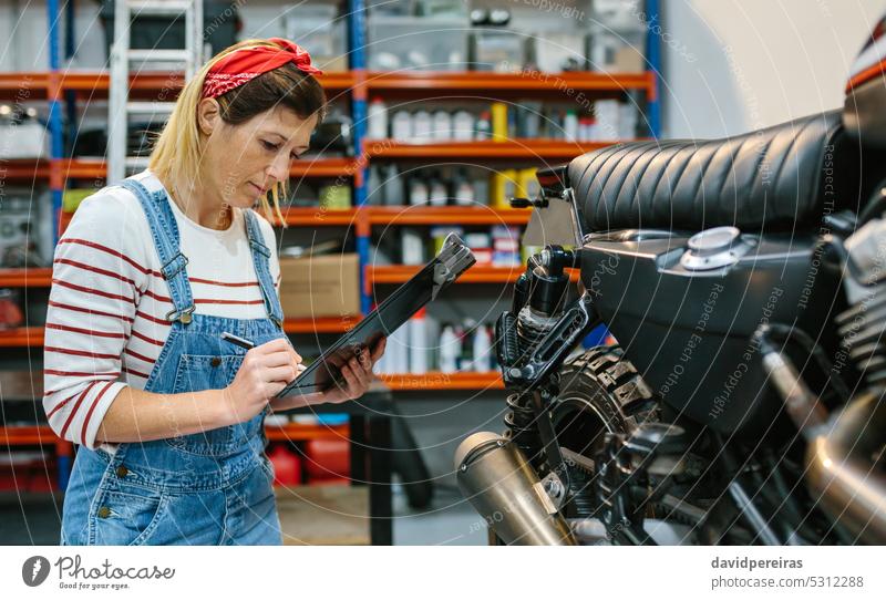 Mechanikerin überprüft Motorrad in der Fabrik Frau Überprüfung Checkliste Prüfung Garage Zwischenablage schreibend Schreibstift Konzentration Versicherung