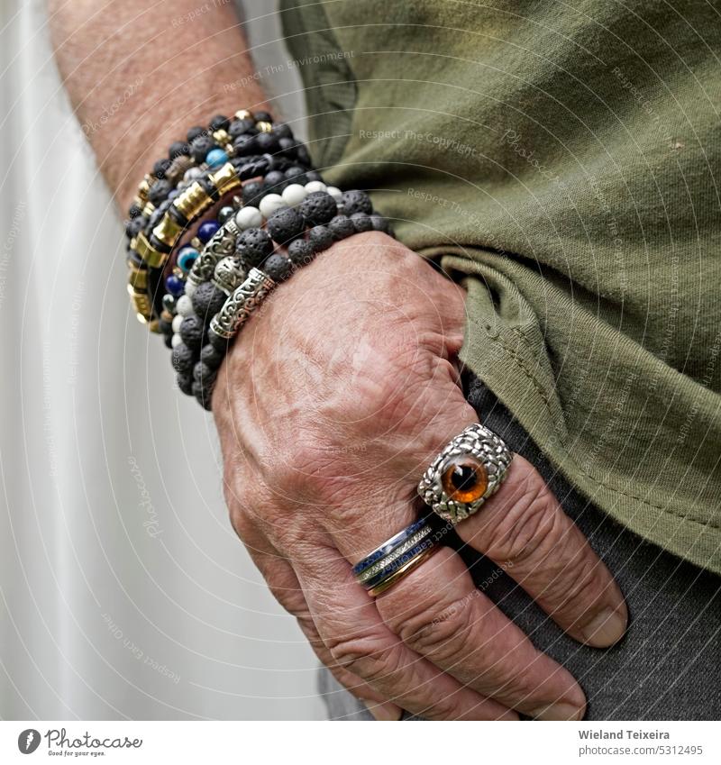 Ein älterer Mann steckt seine Hand in die Tasche seiner grauen Jeans. Er trägt Lavastein und andere panierte Armbänder und besondere Ringe. Person Stein schwarz