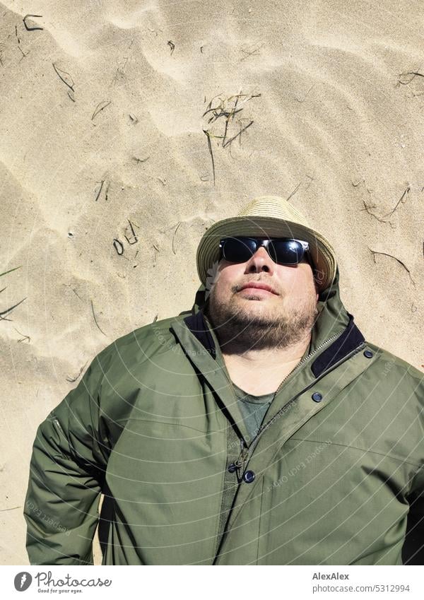 bärtiger Mann mit Strohhut, Sonnenbrille und grüner Wetterjacke liegt im Dünensand an einem sonnigen Tag am Meer Urlauber Wanderer Sommer Frühjahr