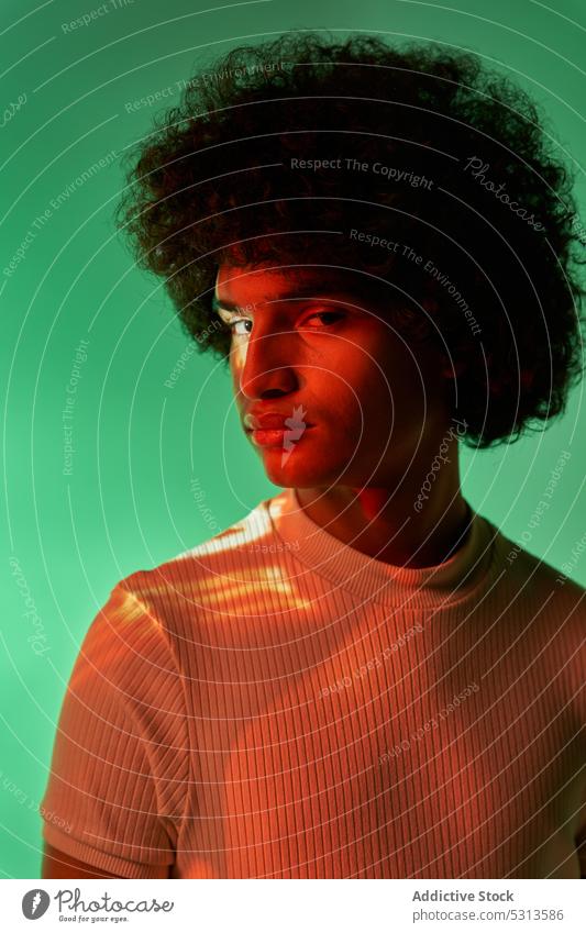 Hispanischer Mann steht vor bunter Beleuchtung und schaut in die Kamera Model farbenfroh mehrfarbig Afrohaar leuchten Projektor glühen kreativ Studioaufnahme