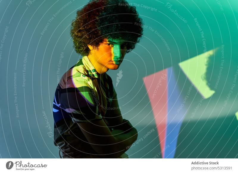 Hispanischer Mann, der vor einer bunten Beleuchtung sitzt und in die Kamera schaut Model farbenfroh mehrfarbig Afrohaar leuchten Projektor glühen kreativ
