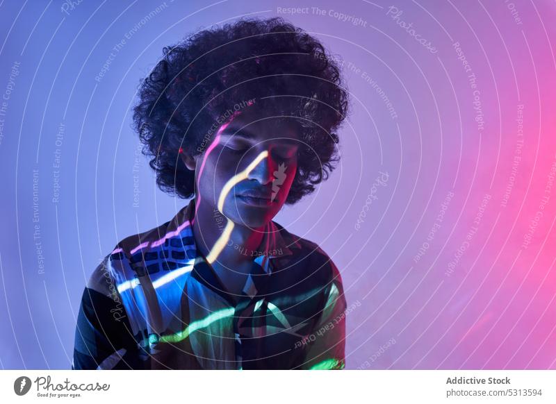Hispanischer Mann vor bunter Beleuchtung, der in die Kamera schaut Model farbenfroh mehrfarbig Afrohaar leuchten Projektor glühen kreativ Porträt Studioaufnahme