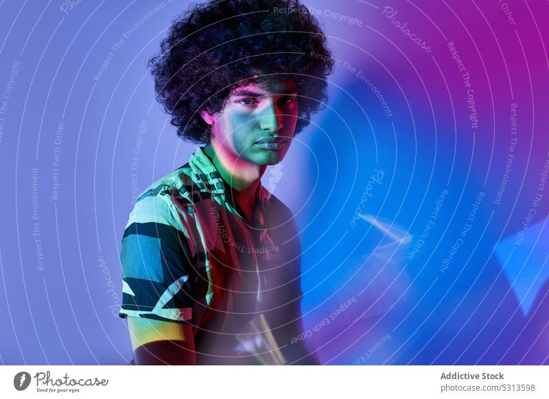 Hispanischer Mann, der vor einer bunten Beleuchtung sitzt und in die Kamera schaut Model farbenfroh mehrfarbig Afrohaar leuchten Projektor glühen Porträt