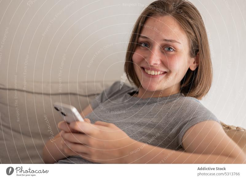 Zufriedene Frau, die auf dem Sofa sitzend mit ihrem Smartphone Nachrichten austauscht benutzend heimwärts Browsen heimisch ruhen Kälte Apparatur Liege Gerät
