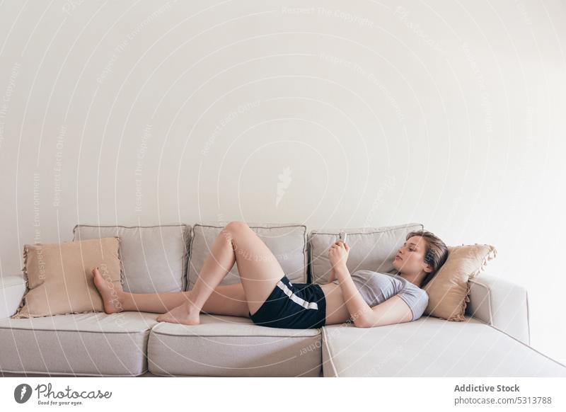 Zufriedene Frau, die auf dem Sofa liegend mit ihrem Smartphone Nachrichten austauscht benutzend heimwärts Browsen heimisch ruhen Lügen Kälte Apparatur Liege