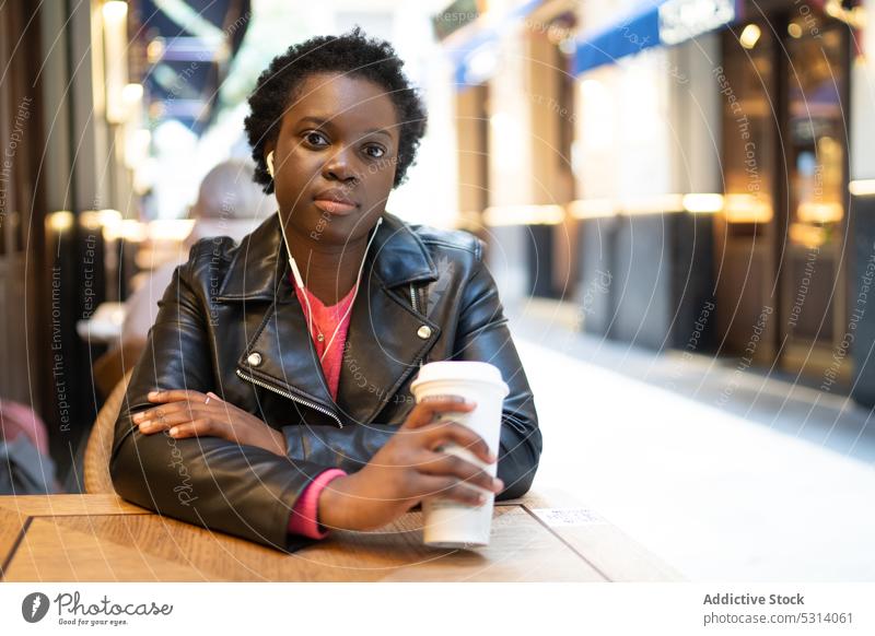 Ernste schwarze Frau mit Kaffee Café Straße trinken Getränk jung Tisch sitzen Erfrischung Afroamerikaner Tasse Kopfhörer Musik zuhören Stil Dame Kantine Jacke