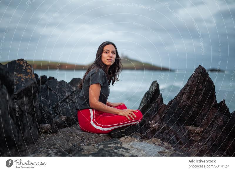 Nachdenkliche junge Frau sitzt auf einem Felsen am Meer Klippe MEER Küste Berge u. Gebirge Natur felsig schäumen Ufer Republik Irland Himmel Glück winken wolkig
