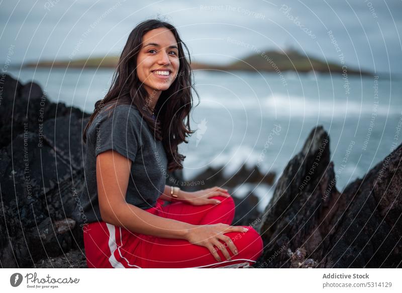 Junge Frau sitzt auf einem Felsen am Meer Klippe MEER Küste Berge u. Gebirge Natur felsig schäumen Ufer Republik Irland Himmel Glück winken wolkig jung Wasser