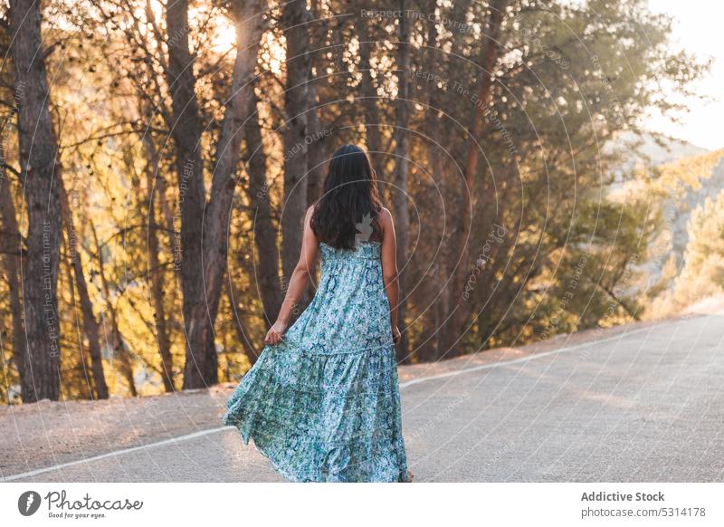Anonyme sorglose Frau, die eine Straße im Wald entlanggeht Reisender Asphalt Sommer Sonnenuntergang Großstadt Spaziergang Baum Urlaub Zaragoza Spanien Kleid