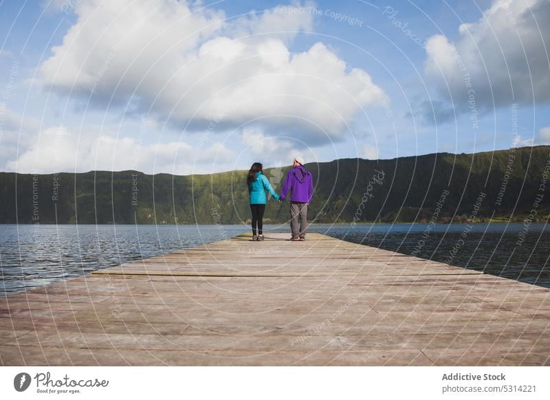 Paar, das an einem bewölkten Tag am Flussufer spazieren geht Pier See bewundern Berge u. Gebirge Händchenhalten Landschaft hölzern Partnerschaft malerisch Natur