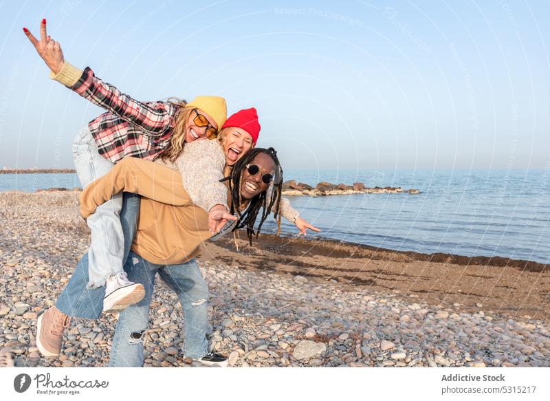 Glückliche Gruppe verschiedener Freunde, die sich am Strand am Meer vergnügen Mann Frauen Huckepack v-Zeichen Küste gestikulieren zwei Finger Lächeln