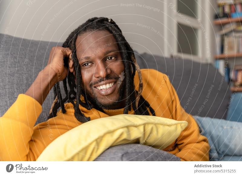 Fröhlicher schwarzer Mann auf Sofa mit Kissen liegend Lächeln positiv heimwärts sich[Akk] entspannen Lügen ruhen Wochenende heiter Kapuzenpulli männlich Glück
