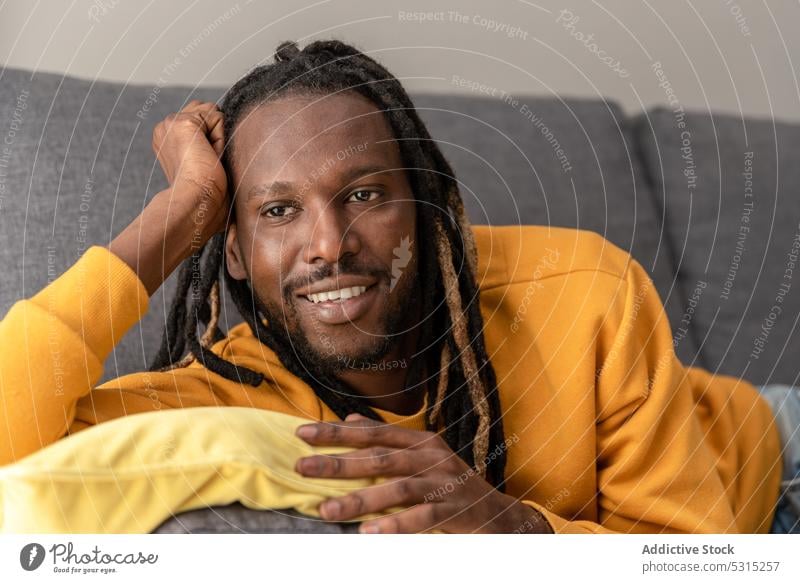 Fröhlicher schwarzer Mann auf Sofa mit Kissen liegend Lächeln positiv heimwärts sich[Akk] entspannen Lügen ruhen Wochenende heiter Kapuzenpulli männlich Glück