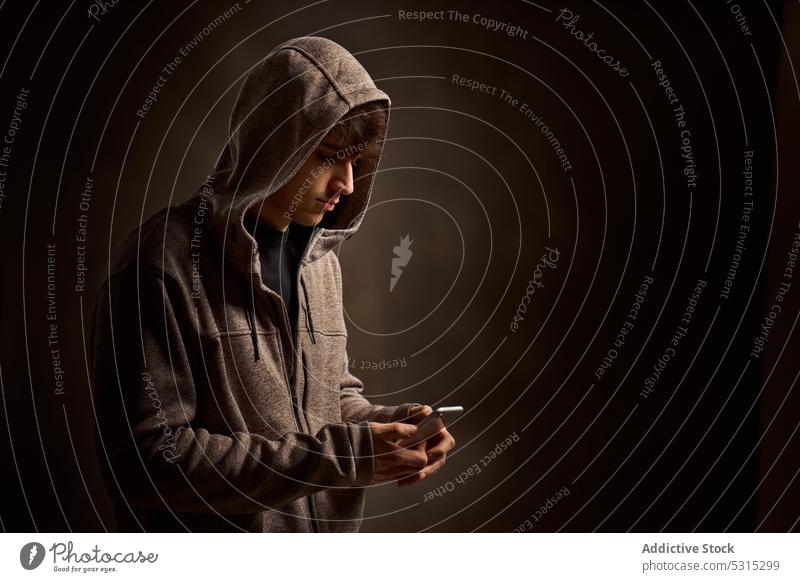 Konzentrierter Mann mit Kapuze, der in einem dunklen Studio auf seinem Smartphone surft benutzend Nachricht Kapuzenpulli Fokus Browsen ernst dunkel Apparatur