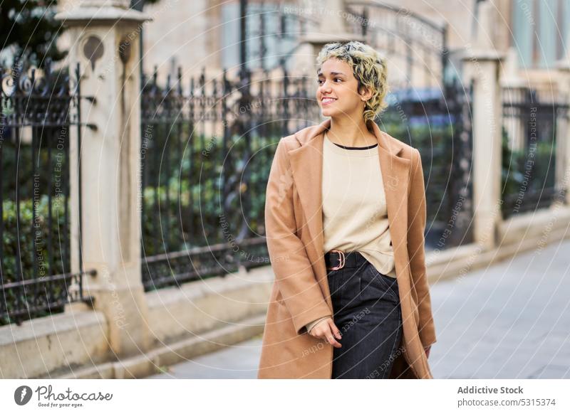 Fröhliche Frau in stilvollem Outfit geht auf dem Bürgersteig Lächeln Spaziergang Gebäude Straße Straßenbelag historisch Stil Großstadt positiv blond trendy jung