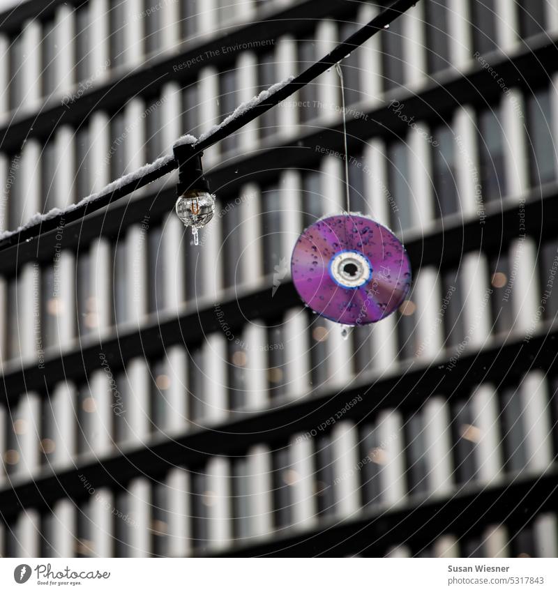 Lichterkette und lilafarbene CD mit Schneeresten und Eiszapfen vor Bürohausfront. Erinnerung an Sommer. Lichterkette mit Eiszapfen cd-rom CD mit Wasertropfen