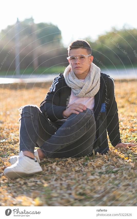 Porträt eines Teenagers mit Brille, der in einem Park auf dem Boden sitzt und in die Kamera schaut. männlich Straße jung Mann Junge Jugend Freizeitkleidung