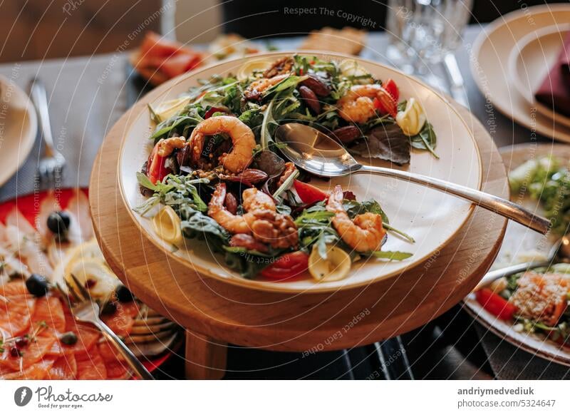 Close up festlichen Tisch mit frischem Salat mit Grill Meeresfrüchte, Grüns und Tomaten auf dem Teller auf Holzständer. Bankett Tisch im Restaurant mit Snacks, Käse Vorspeisen und Salami, Urlaub Catering