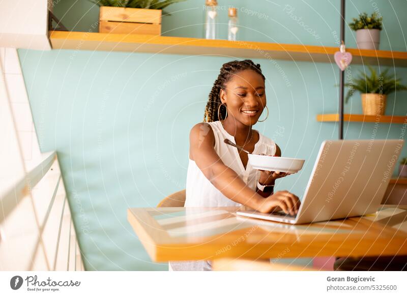 Junge schwarze Frau, die ein gesundes Frühstück zu sich nimmt, während sie in einem Café am Laptop arbeitet Afrikanisch Afroamerikaner Afro-Look Afrofrisur