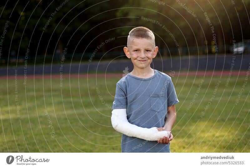 Lächelnder kleiner Junge mit gebrochener Hand schaut in die Kamera draußen auf dem Sportplatz am Sommertag. 9 Jahre Kind hält seine gesunde Hand eine andere gebrochene Gliedmaße mit Gipsverband auf dem Arm. Gesundheit Konzept