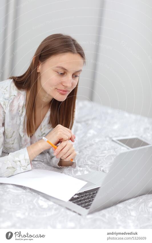 Schöne junge Frau arbeitet, mit Laptop-Computer und macht Notizen im Schlafzimmer zu Hause. Freiberuflerin. Schreibt, tippt. Mädchen überprüft soziale Apps. Kommunikation und Technologie Konzept