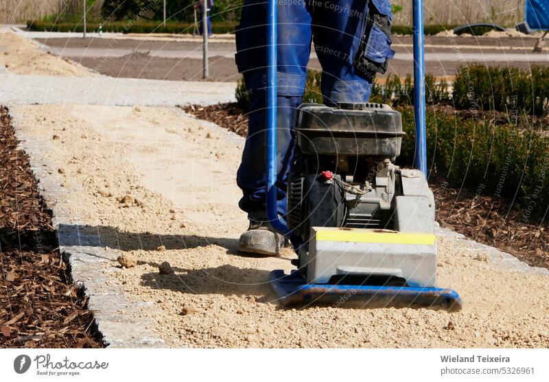Beine und Füße eines Mannes, der eine Sandverdichtungsmaschine bedient. Der Mann rüttelt den Sand für einen neuen Weg. blau Boden im Freien Menschen männlich