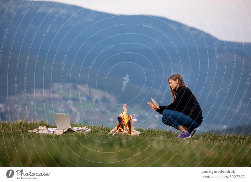 junge Frau Freiberufler sitzt am Feuer und wärmt sich die Hände in den Bergen am Abend. Touristisches Mädchen ruht sich aus und arbeitet am Laptop im Freien