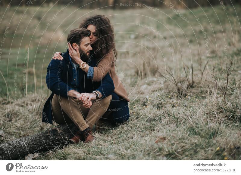Glückliche kaukasische Liebhaber sitzen am Ufer des Sees. Junges Paar umarmt sich an einem Herbsttag im Freien. Ein bärtiger Mann und eine lockige Frau in der Liebe. Valentinstag. Konzept der Liebe und Familie.