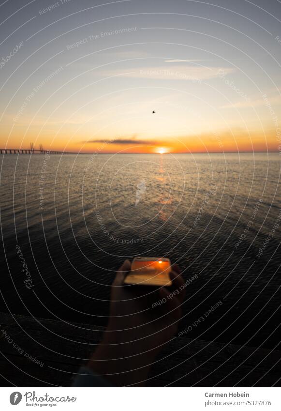 Sonnenuntergang über dem Meer mit Spiegelung im  mit der Hand gehaltenen Handy, links im Bild ist eine Brücke Außenaufnahme Frau Textfreiraum Landschaft Tourist