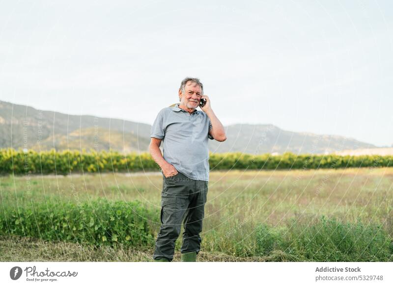 Glücklicher reifer Mann, der im Sonnenlicht auf dem Lande mit seinem Smartphone spricht reden Landwirt Lächeln Landschaft Hügel Feld positiv Natur Ackerbau