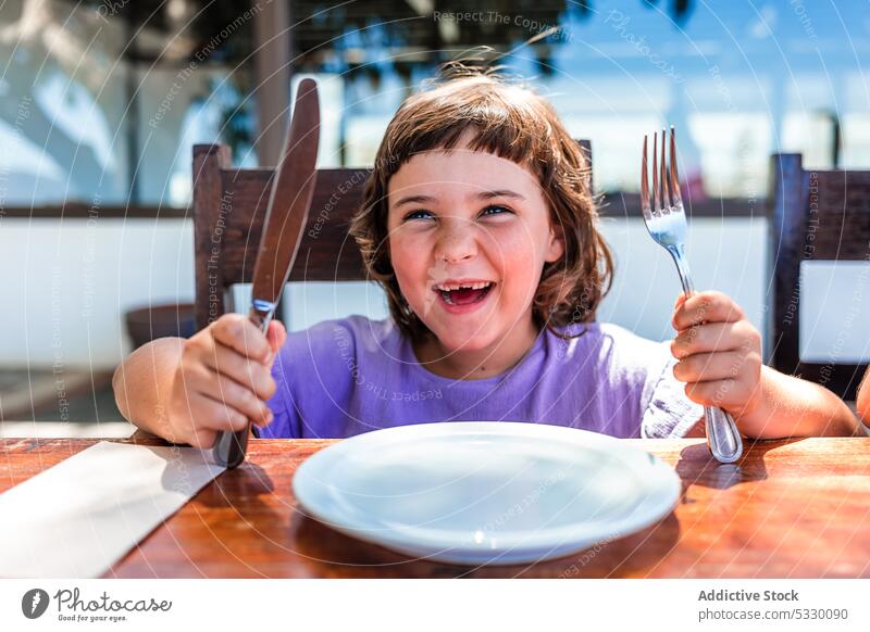 Lustiges kleines Mädchen mit Silberbesteck und Teller im Cafe Café Kind Mund geöffnet warten Tisch hungrig Kindheit Lebensmittel hölzern niedlich Vorschule