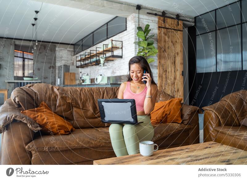 Asiatische Frau spricht mit ihrem Smartphone, während sie an ihrem Laptop arbeitet reden freiberuflich abgelegen Arbeit heimwärts Sofa Anruf Apparatur Gespräch