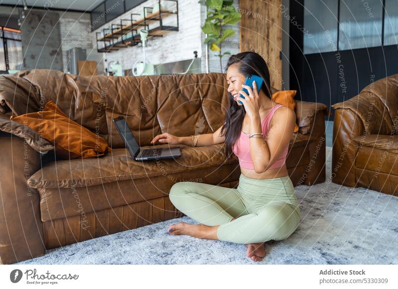 Fröhliche Frau, die auf dem Sofa im Laptop blättert, während sie auf dem Boden telefoniert Smartphone freiberuflich gemütlich Barfuß Browsen Appartement Telefon