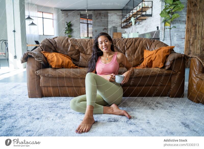 Entspannte Frau trinkt Kaffee auf dem Boden sitzend in einem gemütlichen Wohnzimmer trinken ruhen asiatisch Lächeln Sofa Heißgetränk heimwärts Komfort