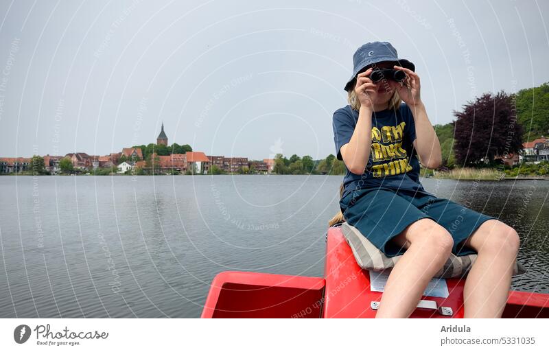 Kind sitzt auf einem Tretboot auf einem See und schaut durch ein Fernglas, Kleinstadt mit Kirchturm im Hintergrund Junge Wasser Stadt Mölln Kirche Ufer Natur