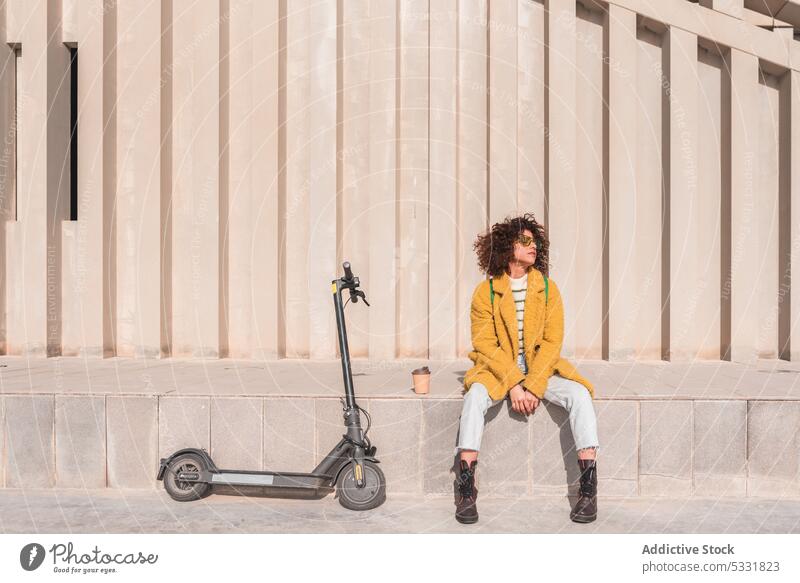 Ruhige Frau sitzt in der Nähe von Elektro-Roller auf der Straße Kaffee elektrisch Tretroller Imbissbude trendy urban Stil Felsüberhang modern Hipster jung