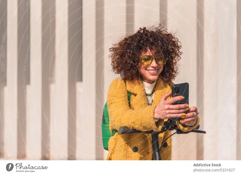 Glückliche Frau, die mit ihrem Smartphone chattet, während sie sich auf einen Motorroller stützt Tretroller elektrisch Stil trendy Lächeln heiter benutzend