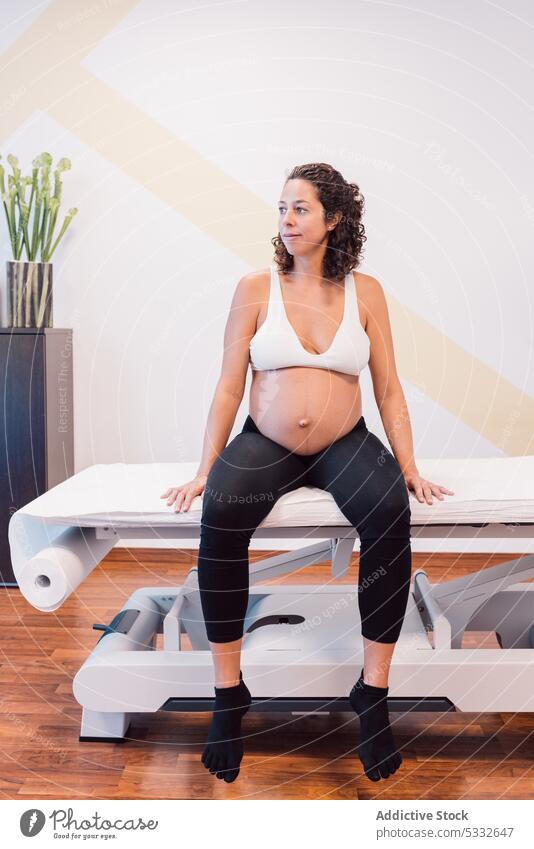 Lächelnde schwangere Frau in Sportkleidung auf einem Massagetisch sitzend geduldig Bank warten Klinik sich[Akk] entspannen Gesundheit erwarten Bauch Glück