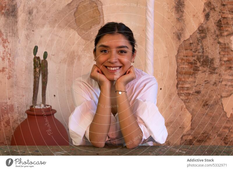 Glückliche Frau sitzt an einem Holztisch im Freien Lächeln heiter Kinn berühren positiv Pflanze Tisch verträumt grün froh jung San Miguel de Allende lässig