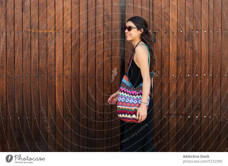 Junge Frau mit bunter Handtasche an einer Holzwand stehend trendy Sonnenbrille hölzern Wand Model Lächeln heiter positiv Glück Stil selbstbewusst schwarz Kleid