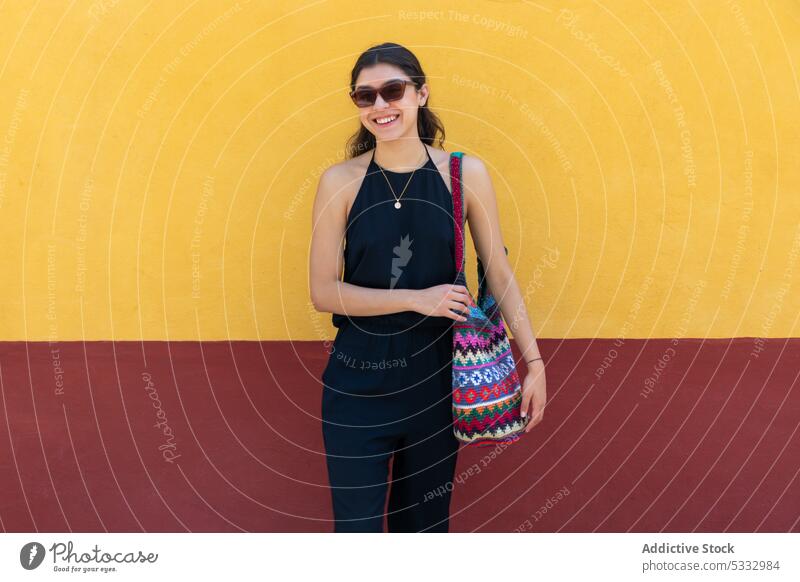 Lächelnde Frau mit Handtasche vor einem bunten Hintergrund Sonnenbrille Mode farbenfroh Wand positiv Stil heiter trendy Farbe hell Outfit schwarz Kleid