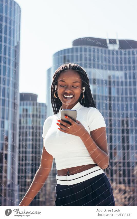 Fröhliche schwarze Frau benutzt Smartphone auf der Straße benutzend cool Kopfhörer Lächeln Browsen positiv Musik Nachricht Glück Surfen Funktelefon heiter